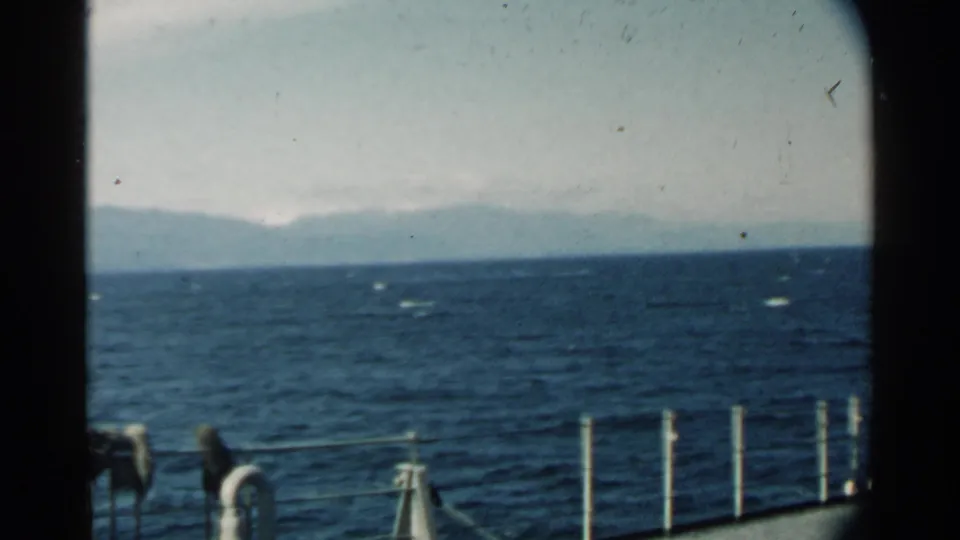 Pearl Harbor-area 8mm frame with foreground guardrail, dark film border, blue ocean, and a faint shoreline in the distance.