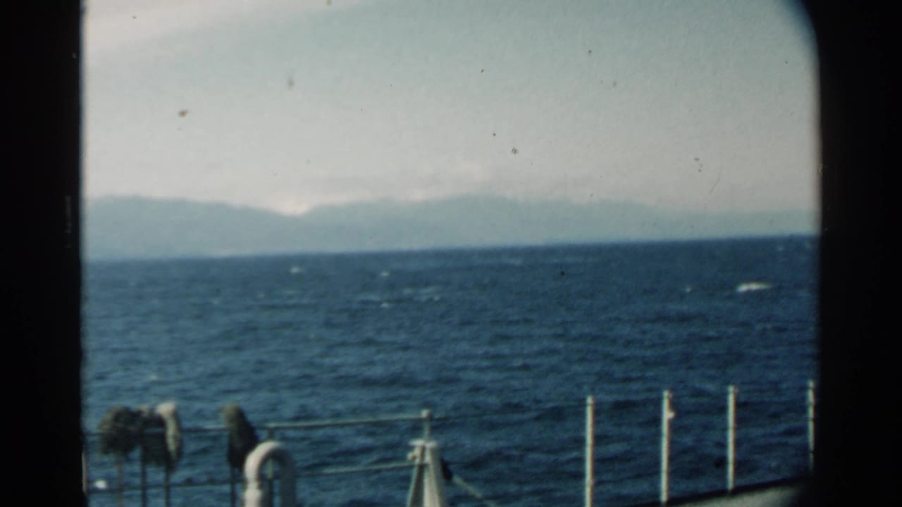 Choppy ocean seen over a boat rail with distant hazy mountains under a pale sky.