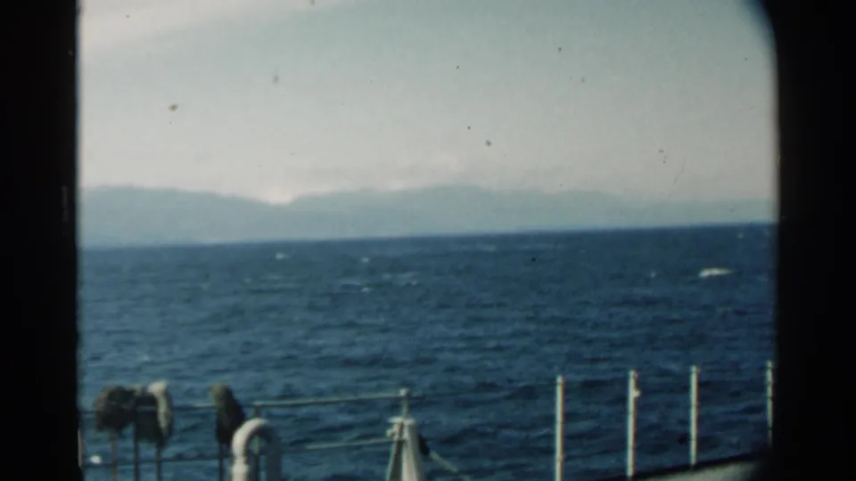 1958 Pearl Harbor home-movie view across blue water toward hazy mountains, with boat railing and hanging gear in the foreground.