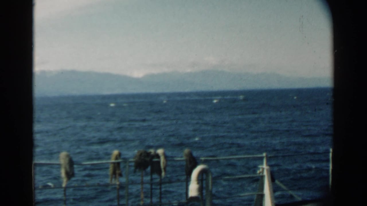 Pearl Harbor water and hazy mountain shoreline appear beyond a boat rail, with small items hanging from the foreground railing.