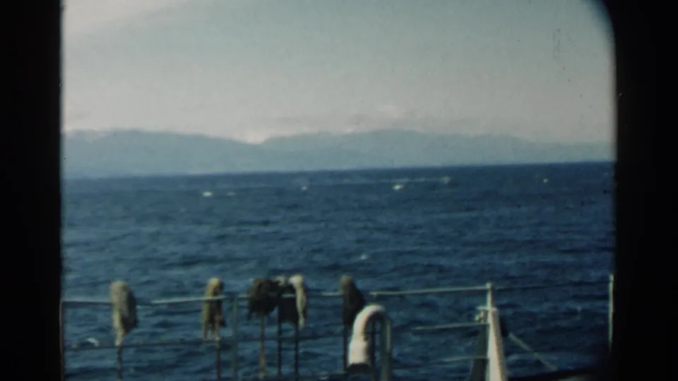 Pearl Harbor water and hazy mountain shoreline appear beyond a boat rail, with small items hanging from the foreground railing.