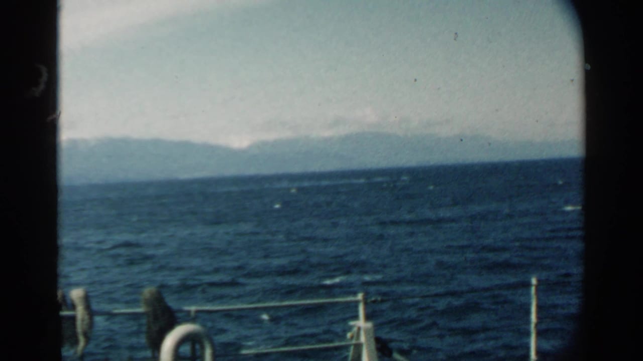 Boat railing in the foreground with ocean waves and a hazy mountain ridge beyond.
