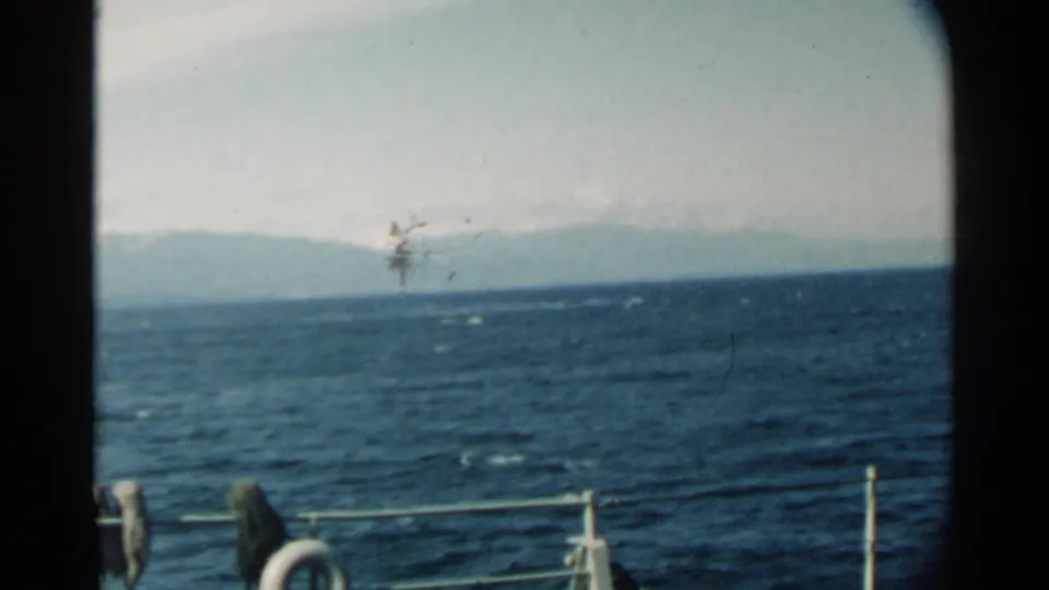1958 Pearl Harbor boat-view frame with choppy blue water, a soft mountain horizon, pale sky, metal railings, and deck equipment in the foreground.