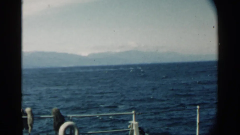 1958 Hawaii home-movie frame from a boat deck, showing railings and gear in front of rippled ocean water and a faint mountain range under haze.