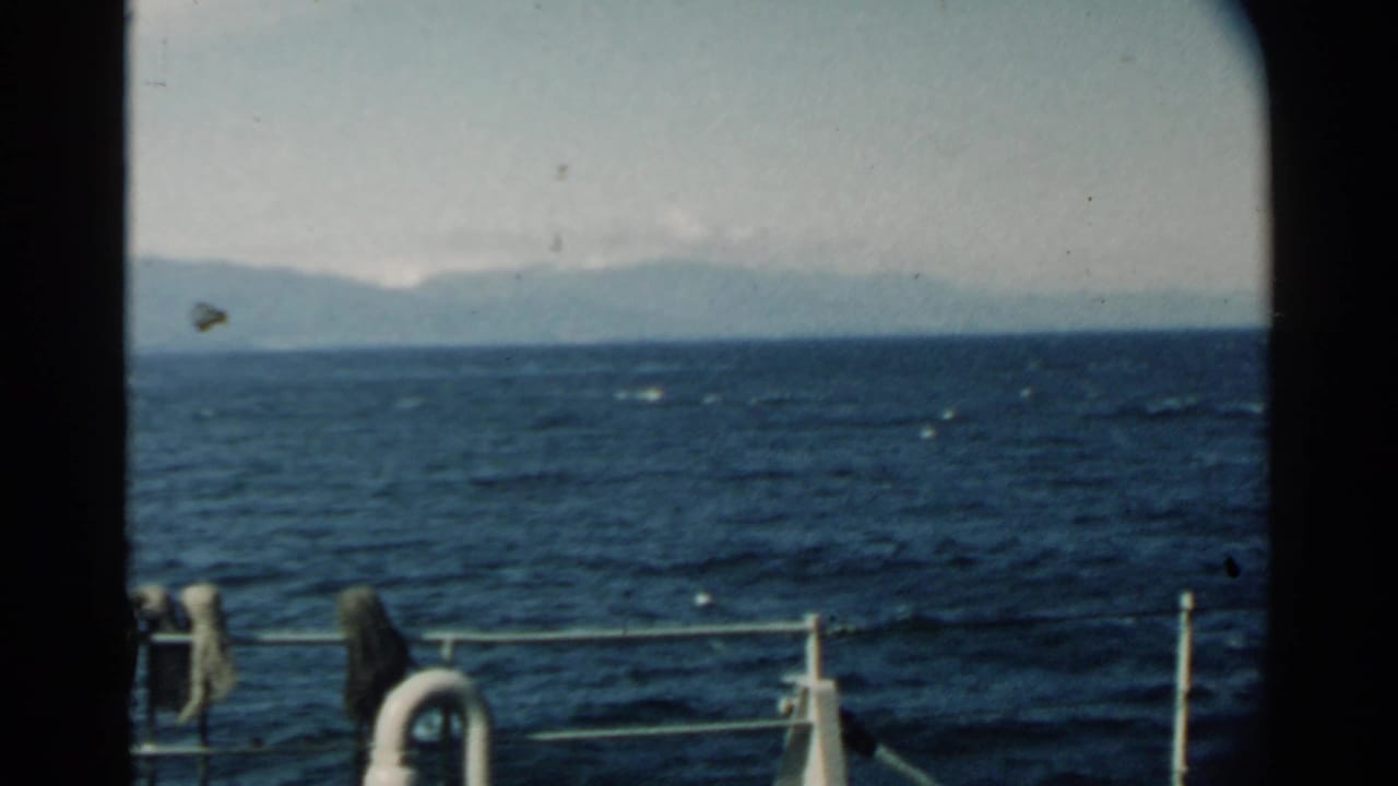1958 Hawaii ship-ride frame with deck railing, white life preserver, choppy blue water, and a faint mountain range beyond the horizon.