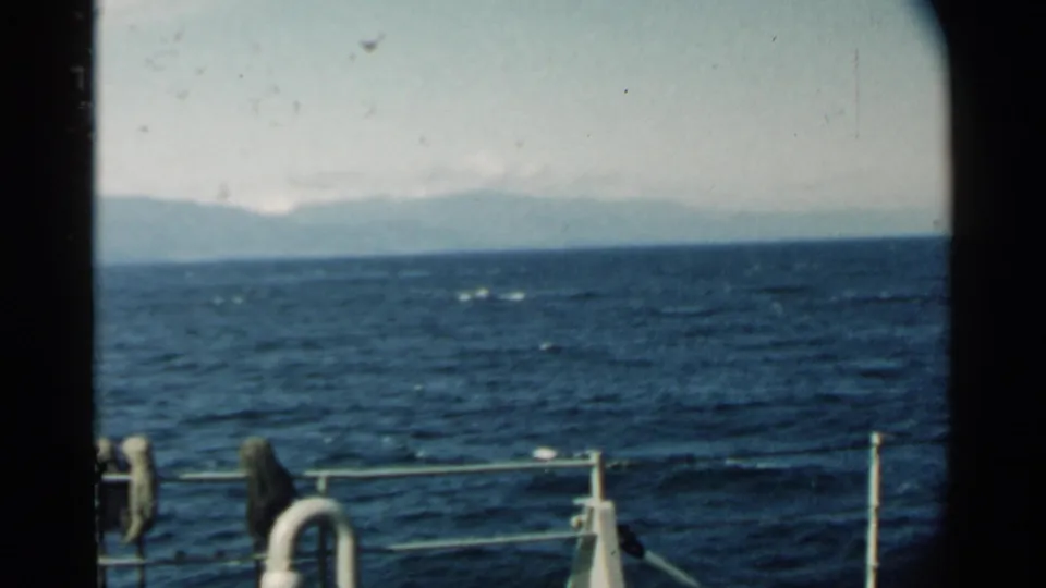 1958 Pearl Harbor-area 8mm still with choppy ocean, hazy shoreline mountains, ship railing, and a white life preserver in the foreground.