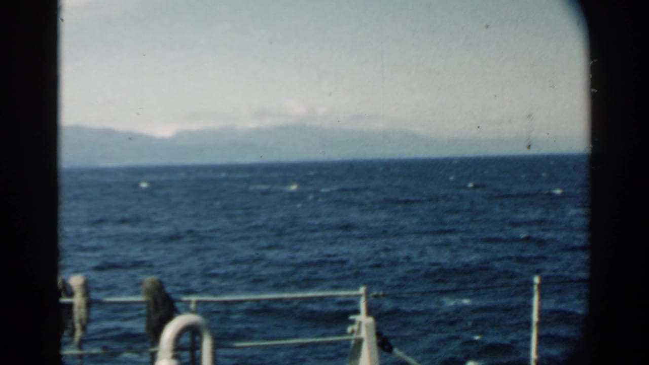Ocean waves seen from a boat with railing in front and hazy mountains on the horizon.