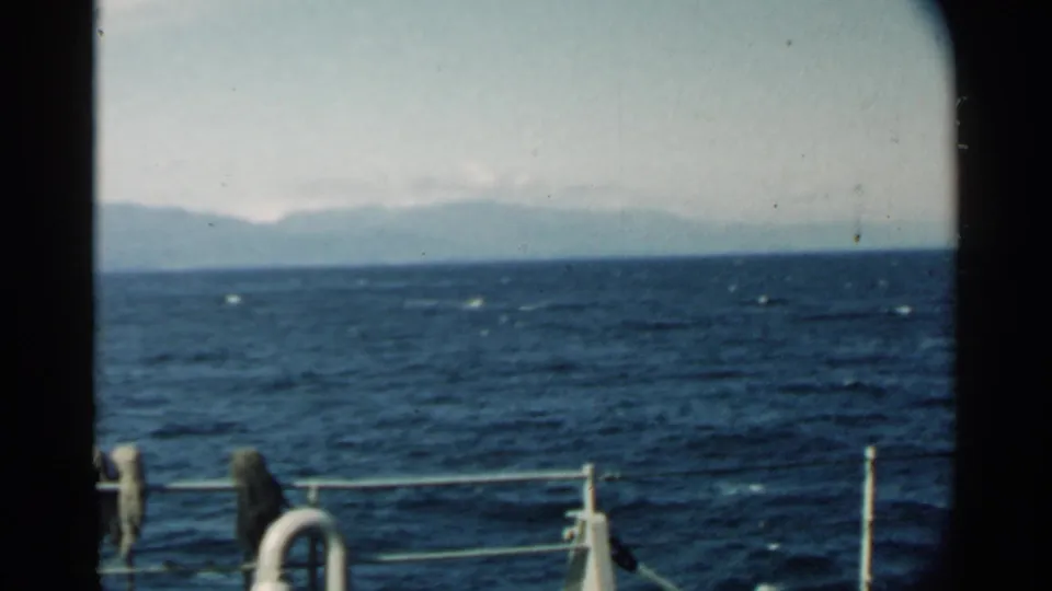 1958 8mm view near Pearl Harbor showing choppy blue water, boat railing, and distant mountainous land under a pale sky.