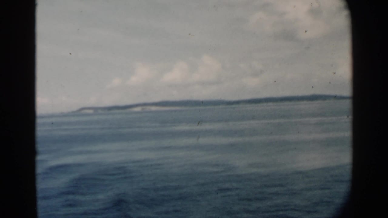 1958 Pearl Harbor-area home-movie frame showing calm blue water, cloudy sky, and a distant island horizon through dark window edges.
