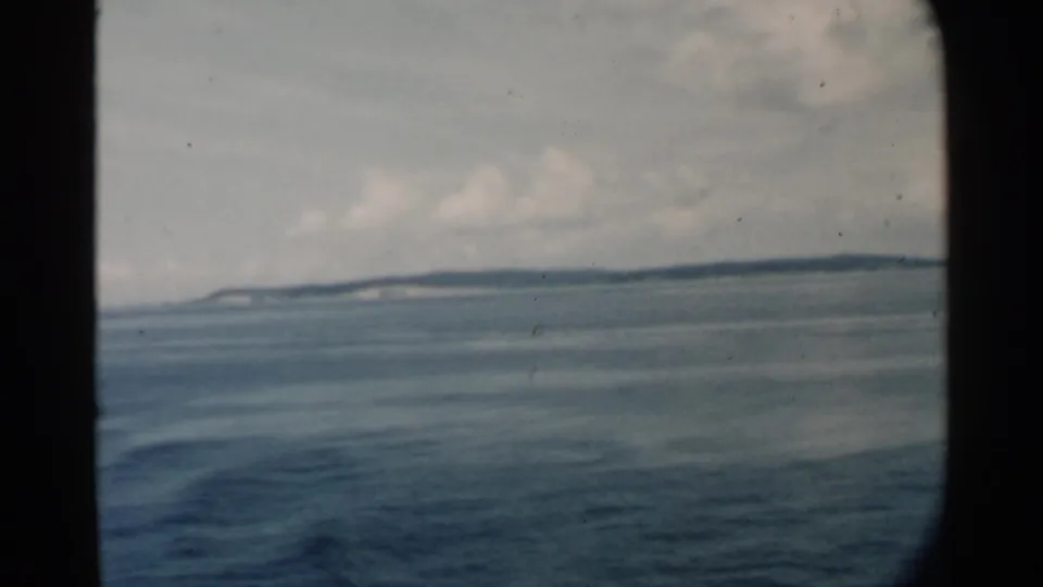 1958 Pearl Harbor-area home-movie frame showing calm blue water, cloudy sky, and a distant island horizon through dark window edges.