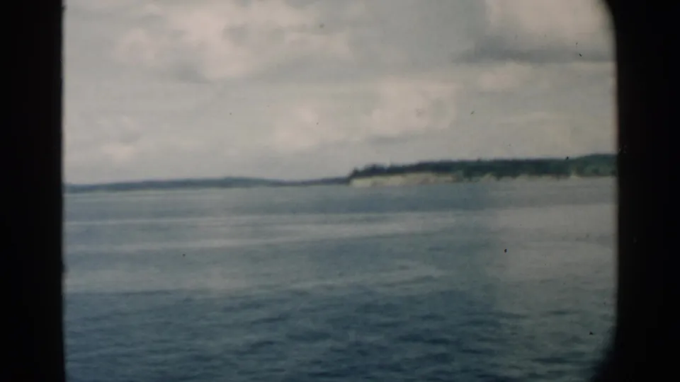 1958 Hawaii home-movie still showing calm water and a distant green shoreline from a vessel, with 8mm frame edges visible.