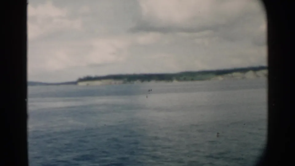 Pearl Harbor-area 8mm frame with a distant shoreline, calm blue water, cloudy sky, and dark vessel-window borders.