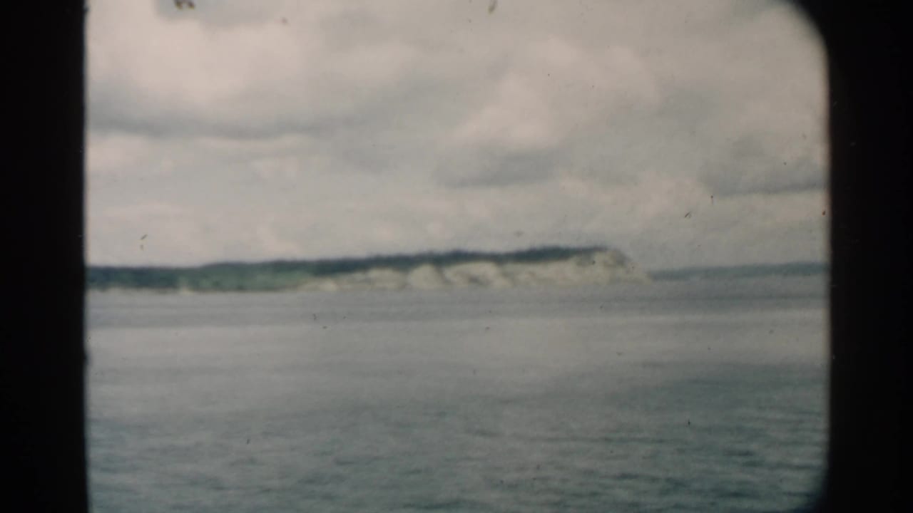 1958 Pearl Harbor home-movie frame showing still water, distant cliffs, and a green island edge below cloud cover.