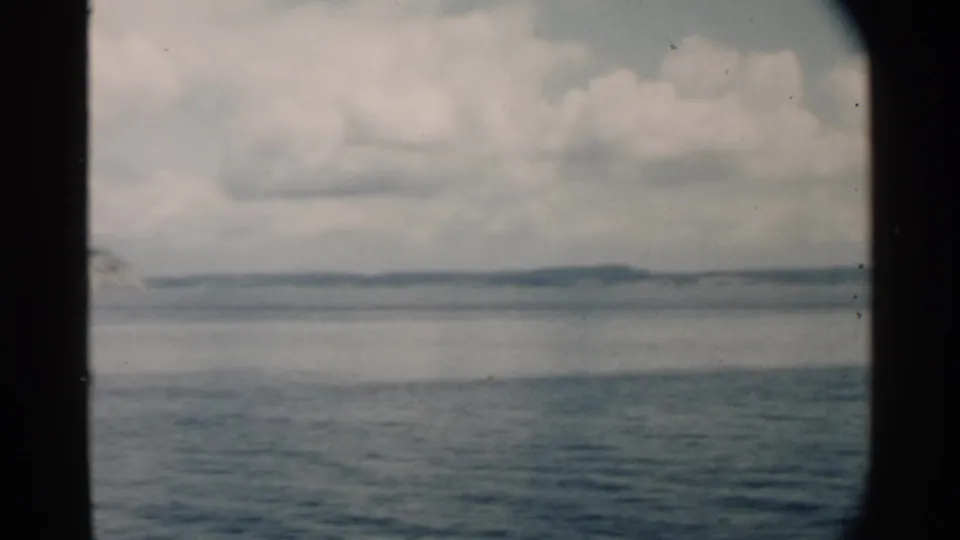 1958 Pearl Harbor, Hawaii home-movie frame showing calm blue water, low distant shore, and cloudy sky through dark window edges.