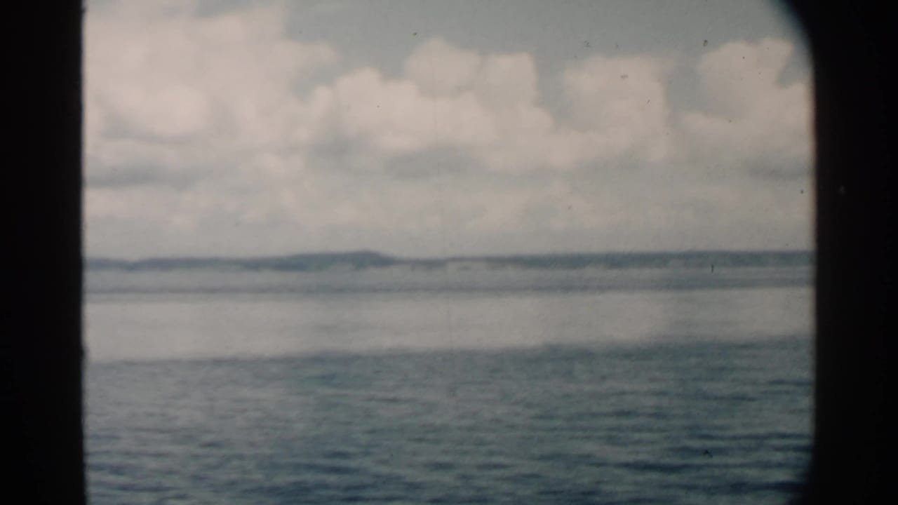 Archival 8mm view from Pearl Harbor metadata, with blue water, pale cloud cover, and a low shoreline framed by a vessel opening.