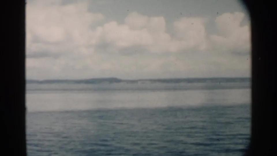 Archival 8mm view from Pearl Harbor metadata, with blue water, pale cloud cover, and a low shoreline framed by a vessel opening.