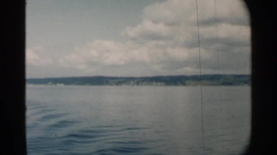 Midcentury 8mm view across Pearl Harbor waters, with a low shoreline, rippled surface, and soft cloud cover visible from a boat.