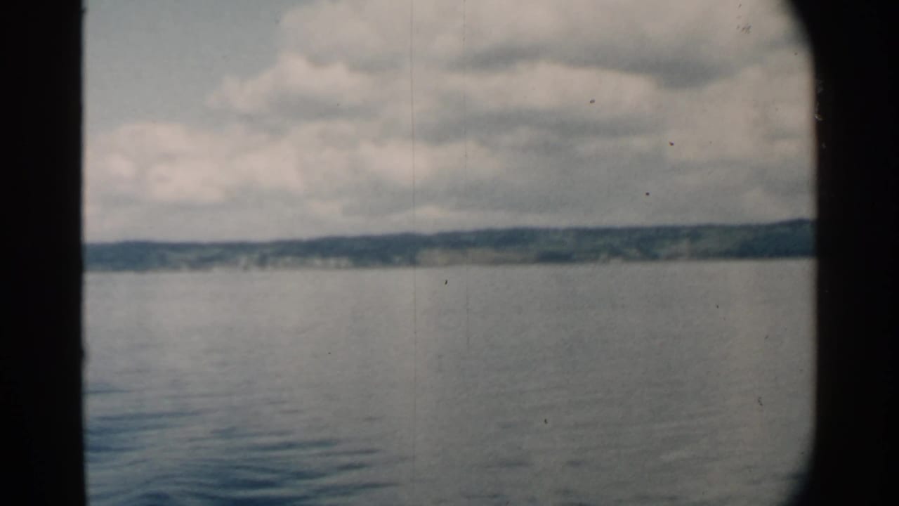 1958 Pearl Harbor home-movie frame looking across calm water toward a low, tree-lined shore beneath heavy clouds.