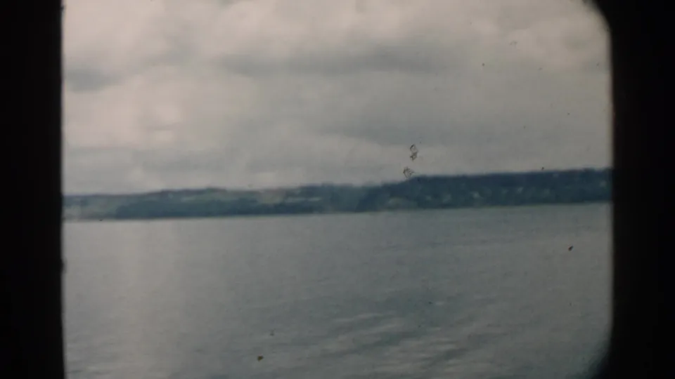 1958 Pearl Harbor-area home-movie frame showing calm blue-gray water, distant shoreline, clouds, and dark window or film edges.