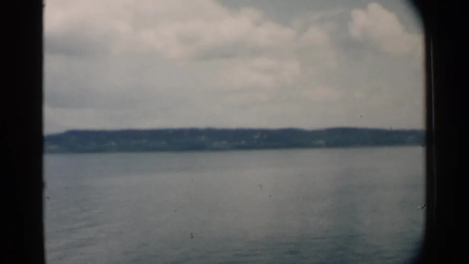 Pearl Harbor-area 8mm frame from 1958 showing blue-gray water, a low far shore, cloudy sky, and slight wake from a moving vessel.