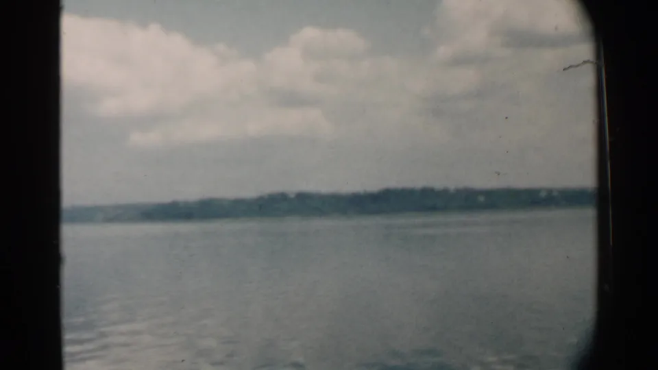 1958 Hawaii home-movie still with calm harbor water, distant shoreline, cloudy sky, visible film grain, and foreground wake patterns.