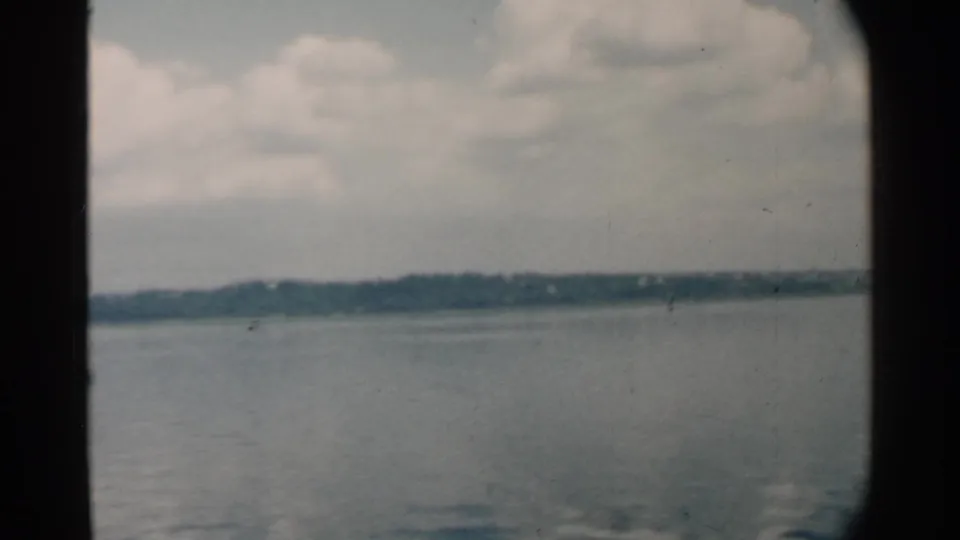 1958 Pearl Harbor home-movie frame showing calm water, a low shoreline, and cloudy sky through a dark vessel window edge.