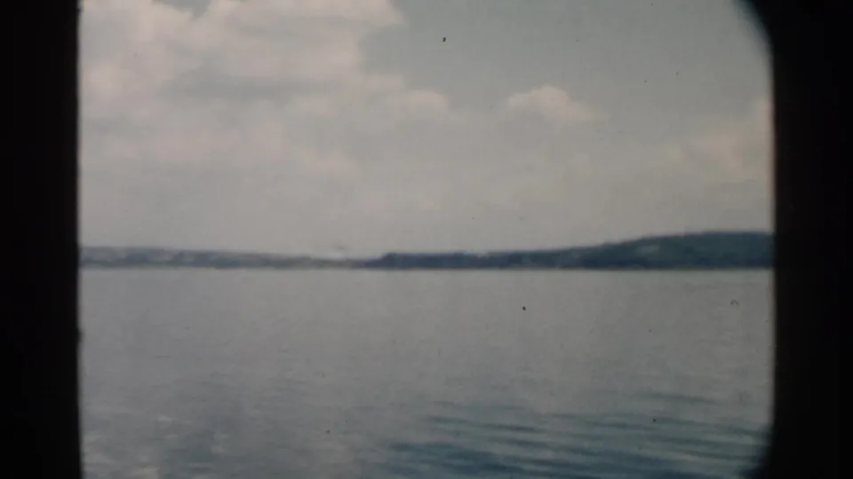 1958 Pearl Harbor home-movie frame showing calm water, a low wooded shore, cloudy sky, and dark boat-window edges.