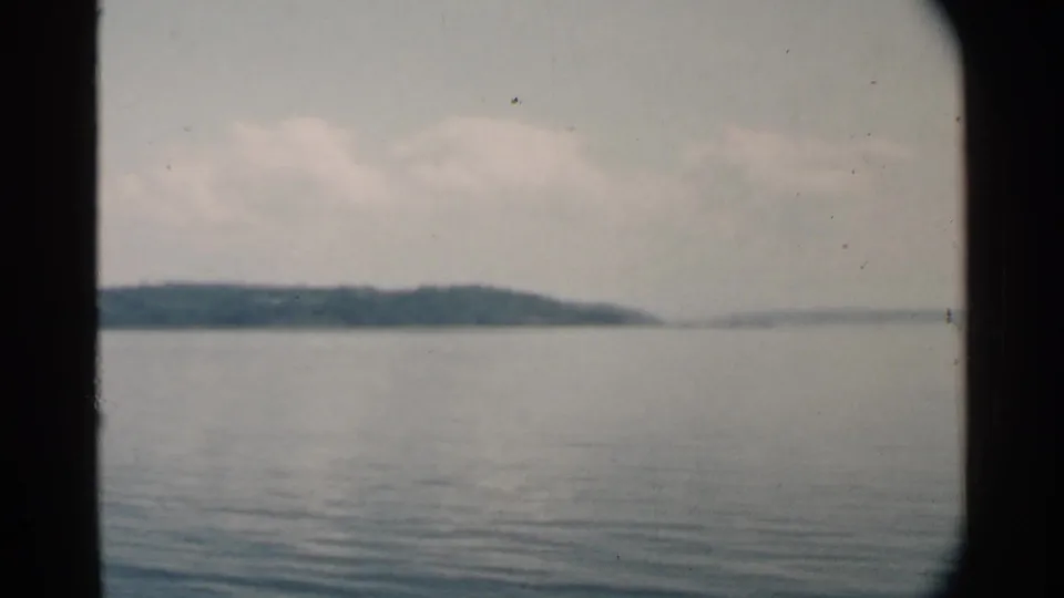1958 Pearl Harbor frame showing calm water, distant wooded land, a soft cloudy sky, and the dark edge of a boat window.