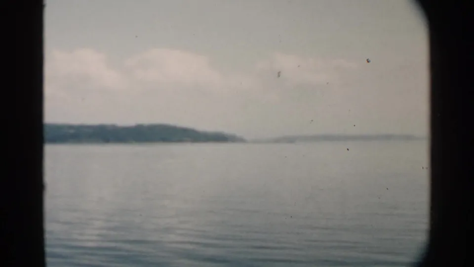 1958 Pearl Harbor home-movie frame showing calm water, hazy shoreline, pale sky, and dark vertical film or window borders.