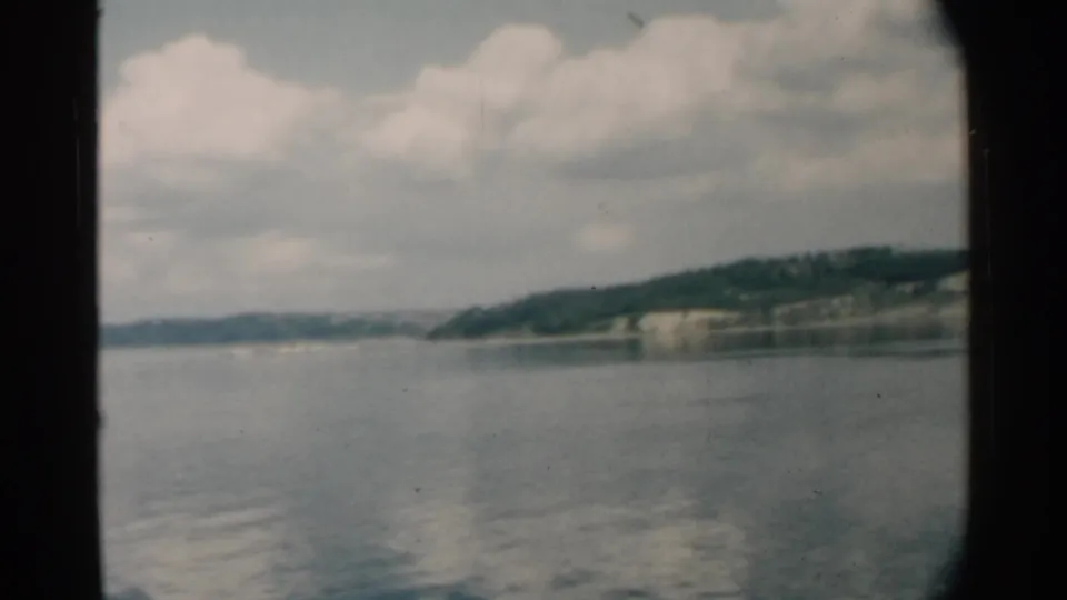 1958 Pearl Harbor frame showing smooth water, a distant wooded shore, pale cliff areas, and broad white clouds.