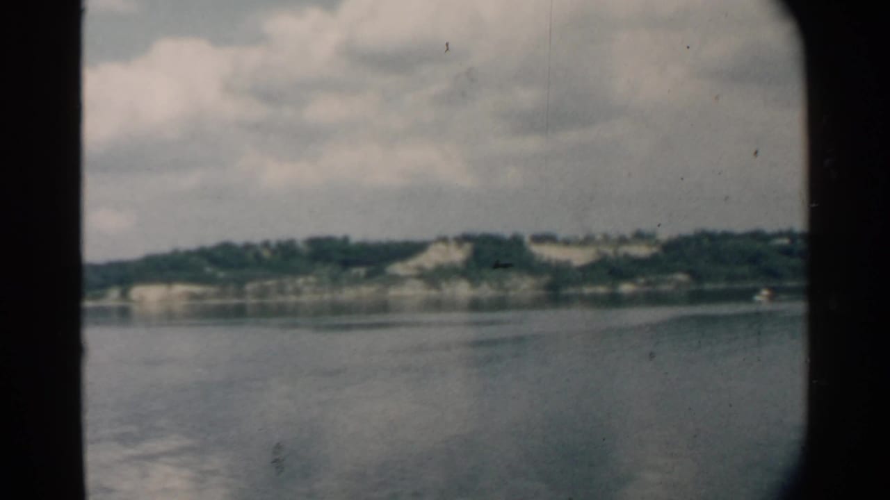 1958 Pearl Harbor home-movie view across calm water toward a green shoreline, framed by a dark rounded window.