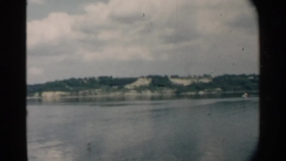 1958 Pearl Harbor home-movie view across calm water toward a green shoreline, framed by a dark rounded window.