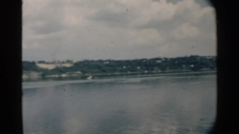 A 1958 Pearl Harbor boat view shows smooth water, green shore, and overcast sky through an 8mm film frame.