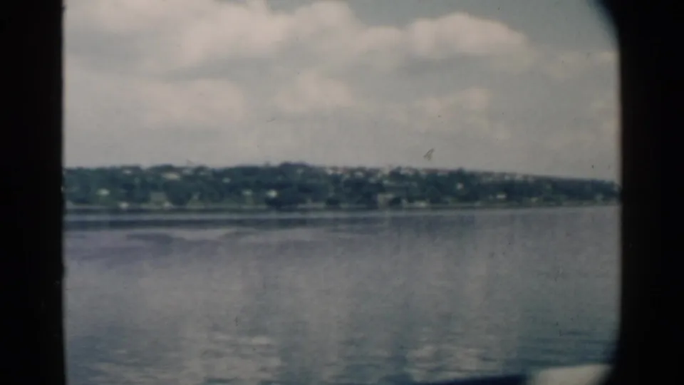 1958 8mm view across calm Pearl Harbor-area water toward a low shoreline under a partly cloudy sky.