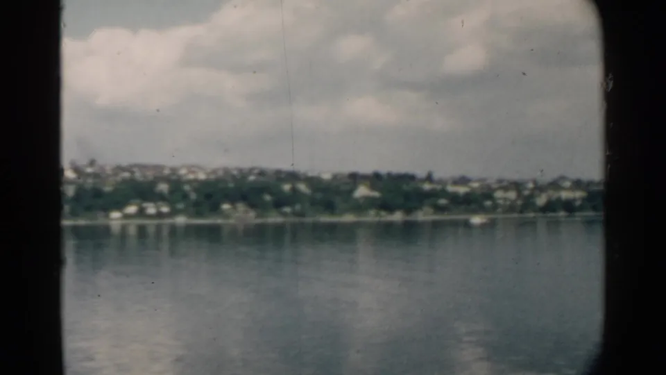 Pearl Harbor, Hawaii, seen from a moving vessel in 1958, with low shoreline buildings and still water under clouds.