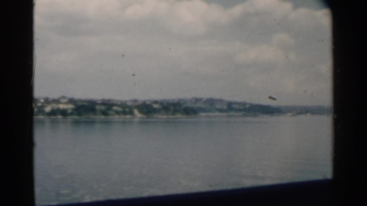 Boat window view across calm water toward a distant shoreline under clouds.