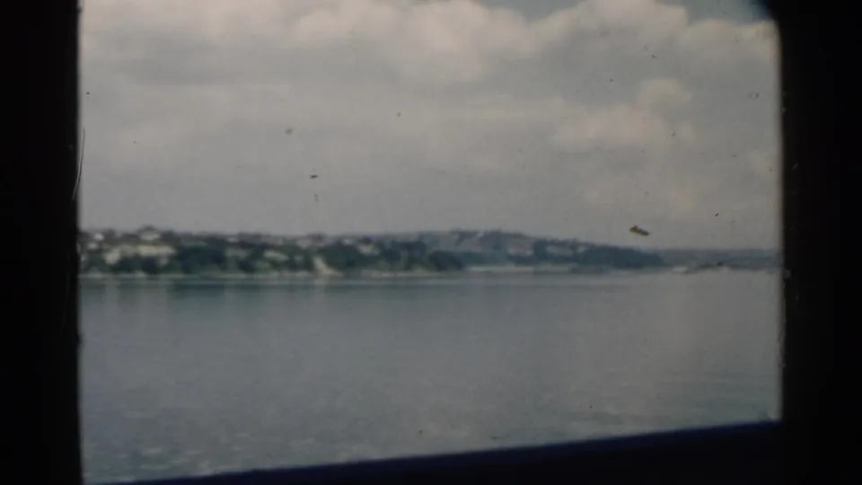 1958 Pearl Harbor home-movie frame looking through a boat window across water toward a distant shoreline and cloudy sky.