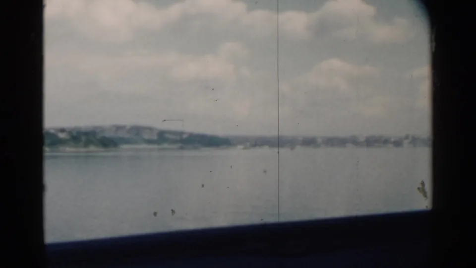 1958 Pearl Harbor view through a boat window, with muted water, distant land, cloudy sky, and visible 8mm film wear.