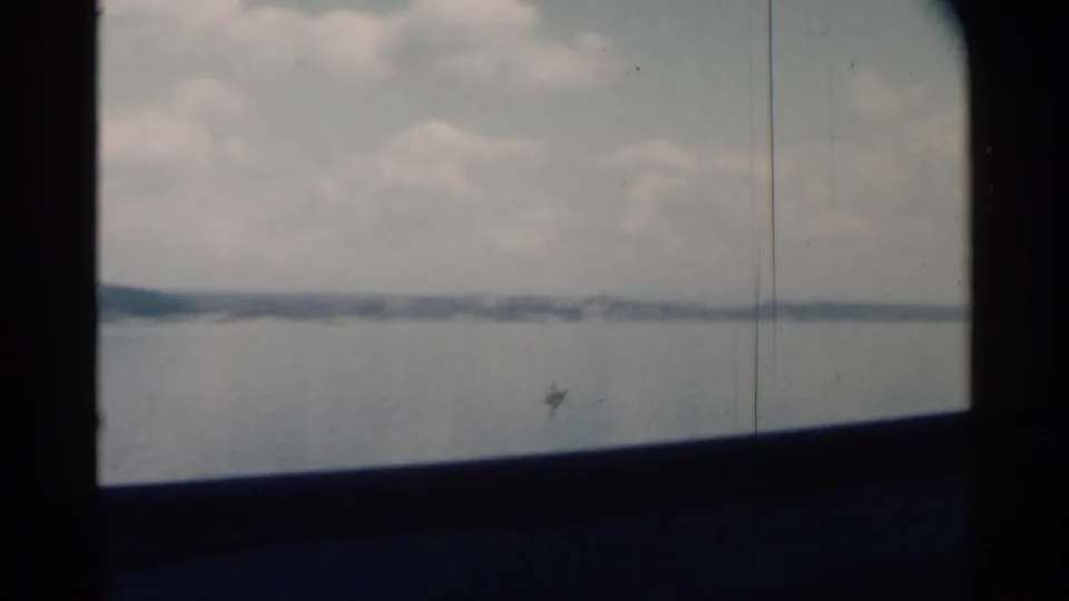 Soft 1958 Pearl Harbor boat-window view with calm water, a low distant shoreline, cloudy sky, and vertical film marks.