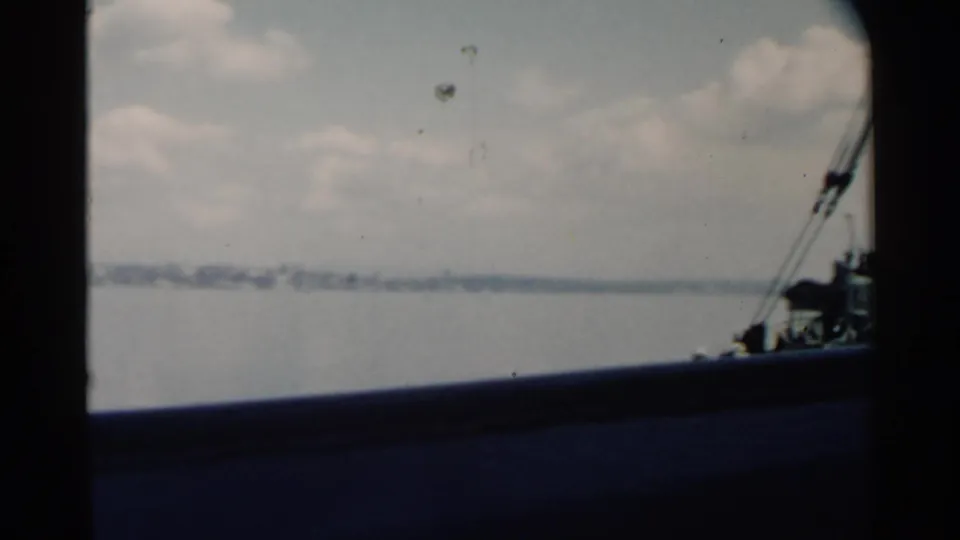 1958 Pearl Harbor home-movie frame showing harbor water, distant shore, cloudy sky, and a small section of boat structure at right.