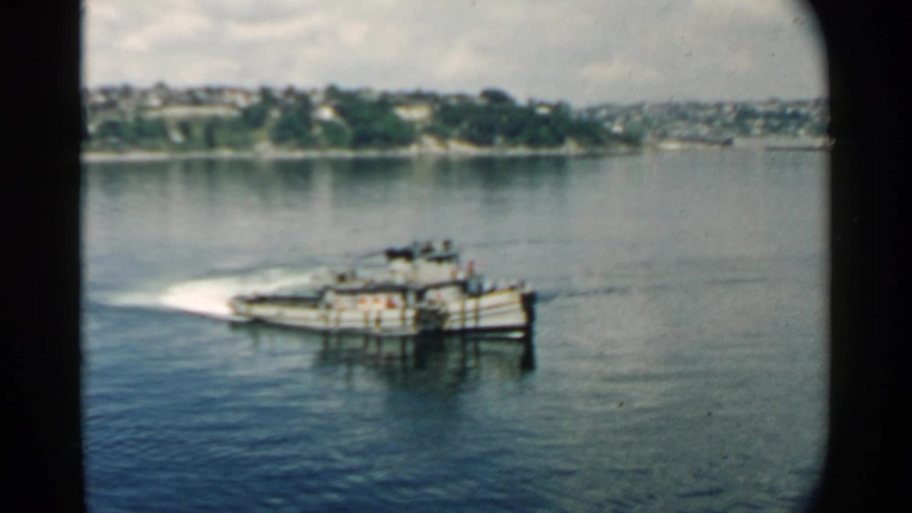 Small working boat crossing calm harbor water with a distant shoreline behind it.