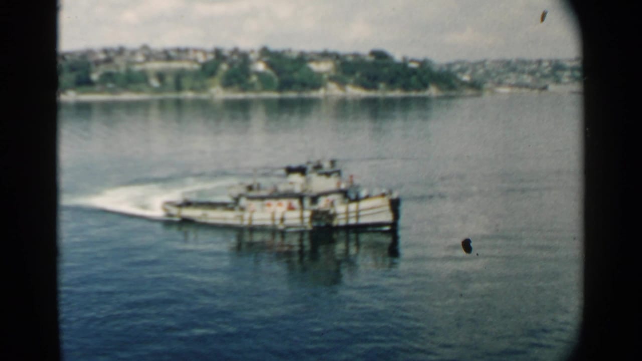 The boat continues across Pearl Harbor-area water in a soft 1958 8mm frame, with shoreline houses or low buildings behind it.