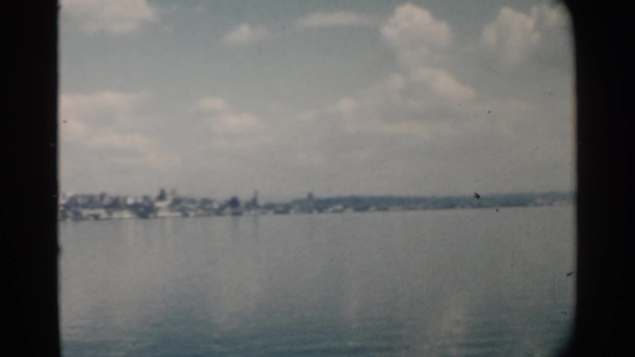 1958 Pearl Harbor home-movie frame showing calm water, a distant low shoreline, clustered buildings, clouds, and dark 8mm frame edges.