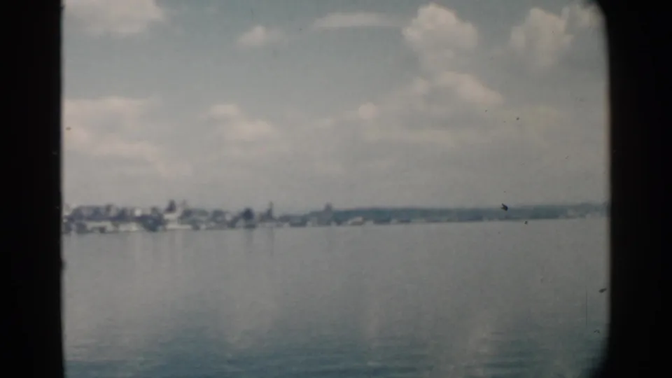 1958 Pearl Harbor home-movie frame showing calm water, a distant low shoreline, clustered buildings, clouds, and dark 8mm frame edges.