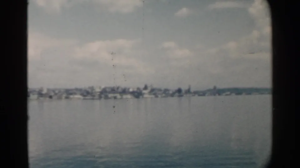 Mid-century 8mm frame from Pearl Harbor showing a low waterfront town line, calm water, soft clouds, and small dust marks on the film.
