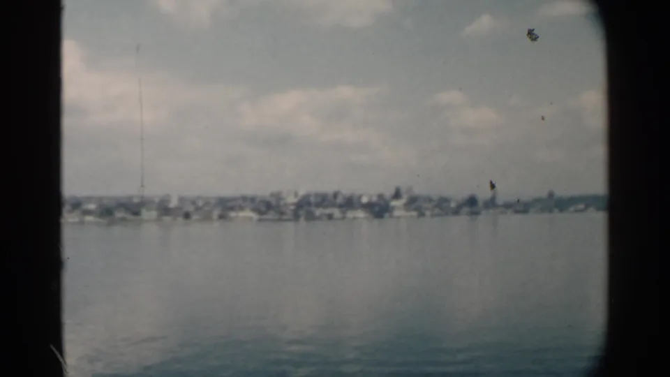 Pearl Harbor home-movie frame with calm foreground water, distant shoreline buildings, cloud cover, and dark 8mm contact-sheet borders.