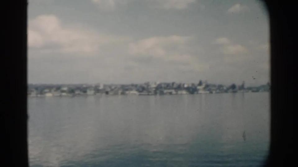 1958 Pearl Harbor home-movie view of distant residential shoreline across still water, framed by dark rounded film edges.
