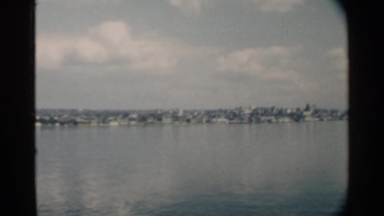 Distant Pearl Harbor shoreline appears through soft 8mm color, with houses and trees beyond calm water in 1958 footage.