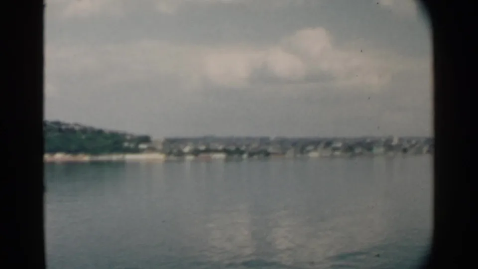1958 8mm view across likely Pearl Harbor water toward a green Hawaiian shoreline and distant buildings.