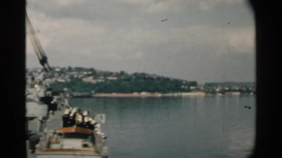 1958 home-movie frame from a boat, with indistinct passengers and a Hawaiian shoreline across the harbor.